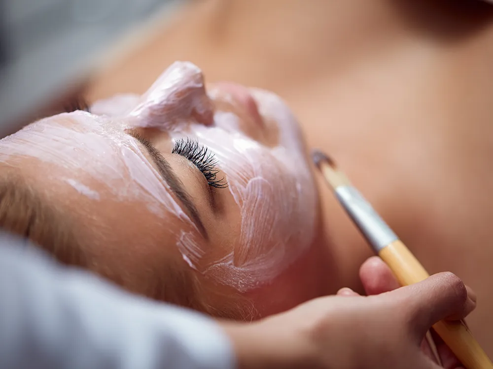 Esthetician brushing a cream mask onto a client's face during a signature facial