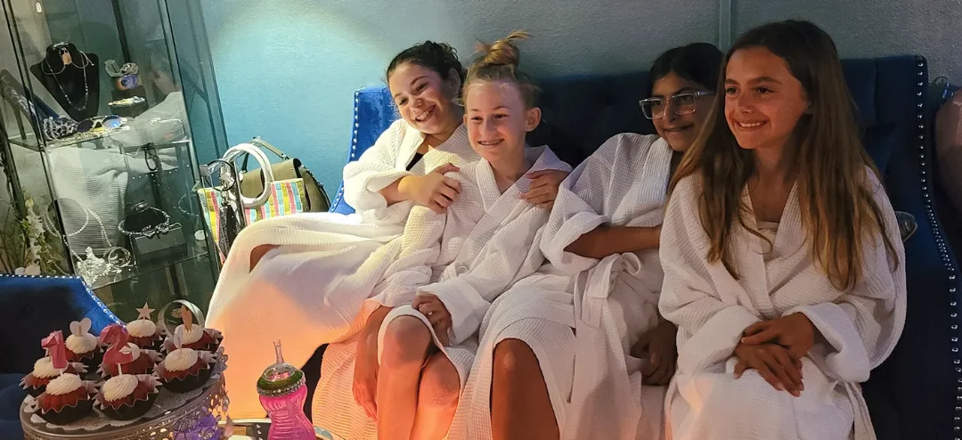 Four girls in white spa robes smiling on a blue velvet sofa beside party cupcakes